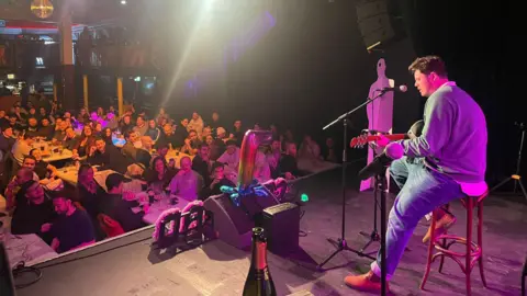 Paddy Merritt Paddy Merritt is seen from the side, sitting onstage at a microphone playing the guitar, with an audience seated at tables.