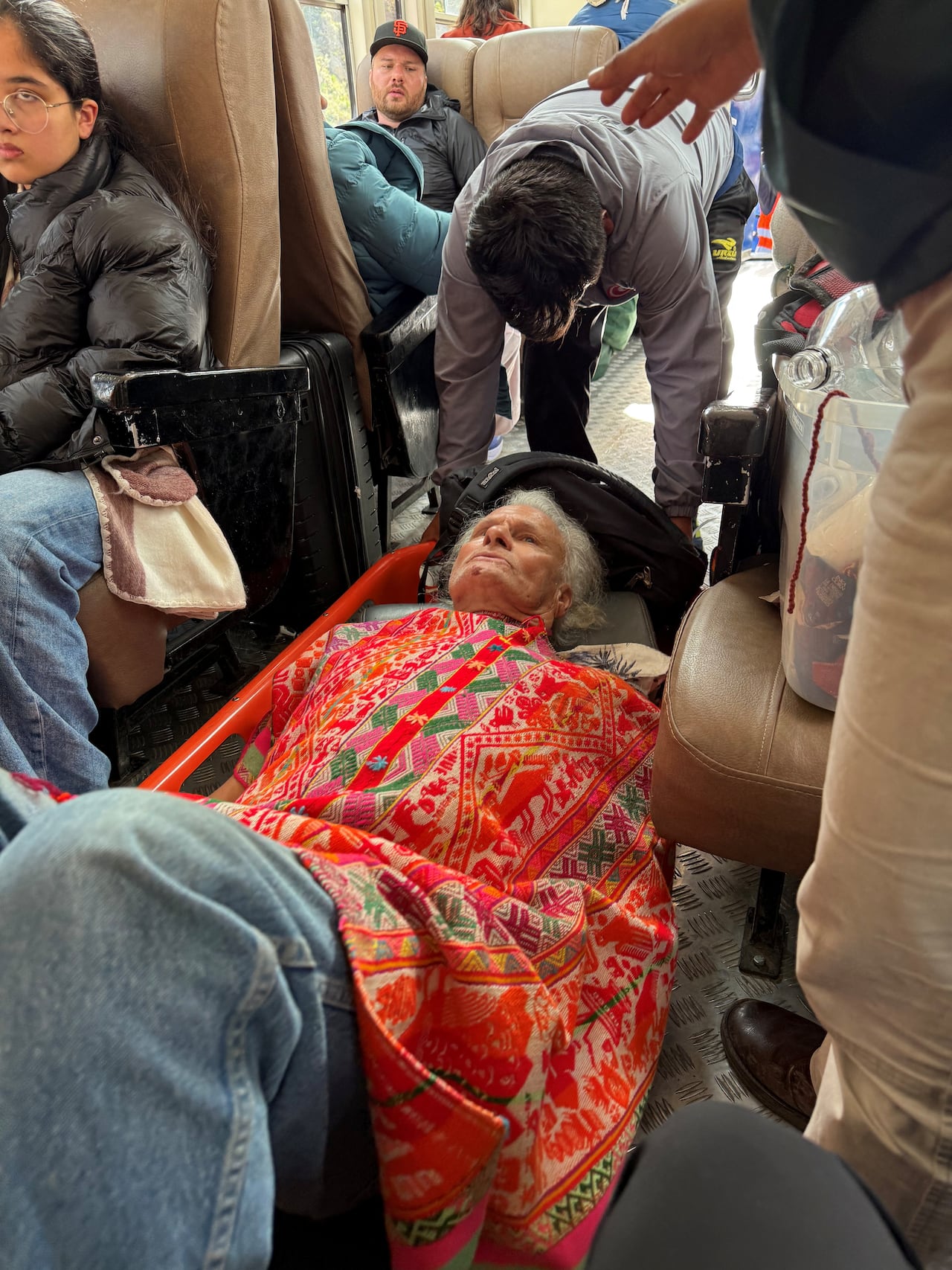 An elderly woman wrapped in a colourful blanket lies on a stretcher in a train.