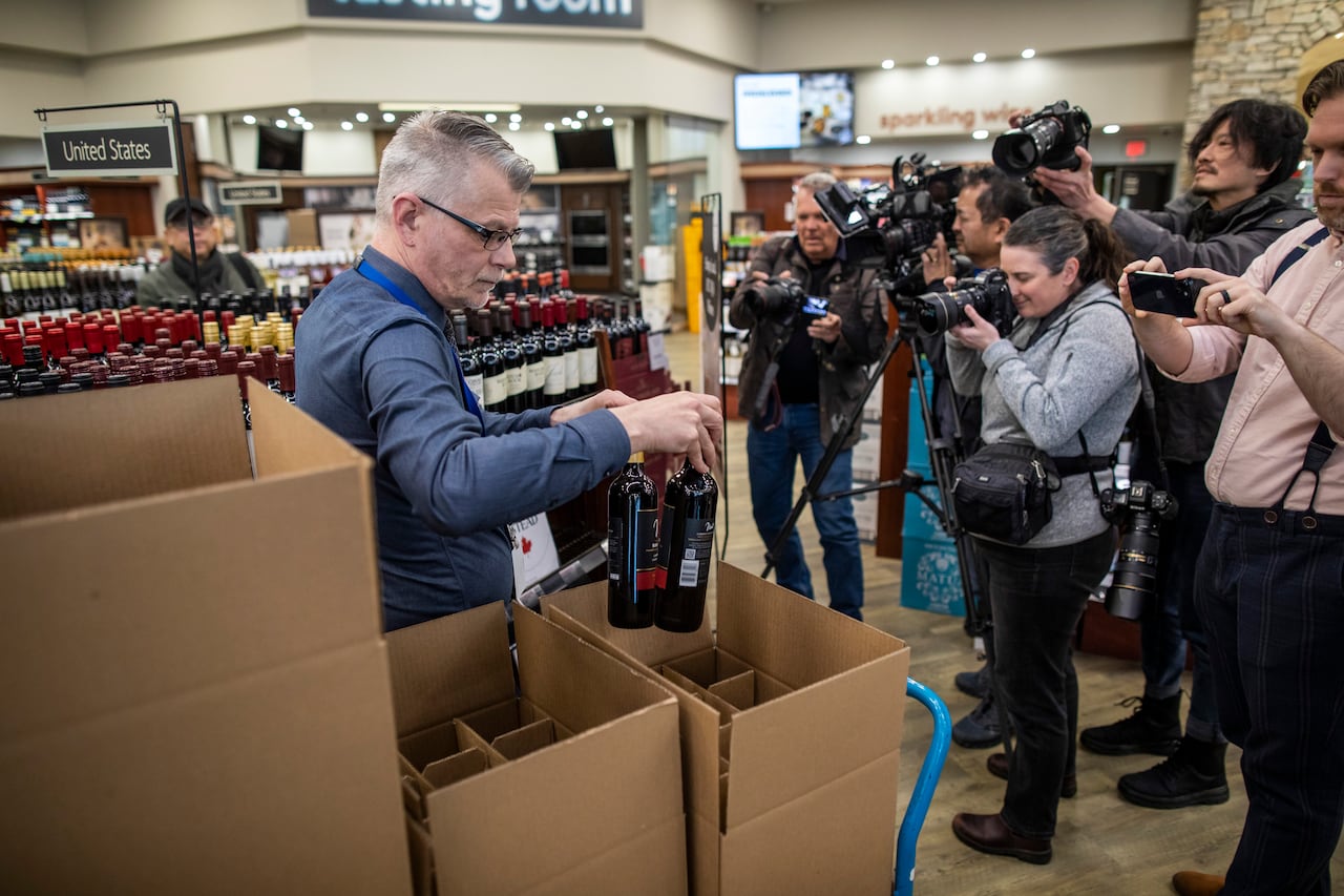 A man holds wine bottles near a box inside of a store while a group of people with cameras take pictures.