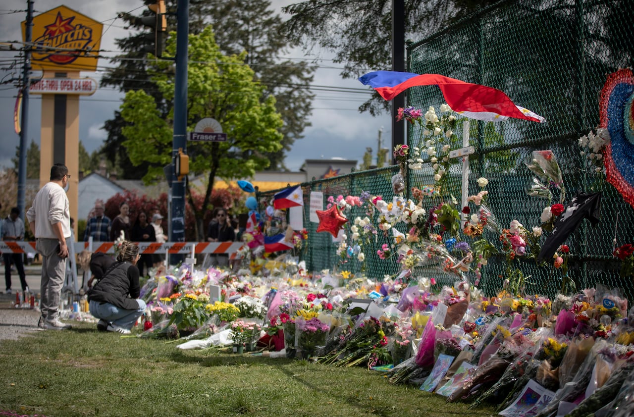 A large pile of flowers and flags sit against a fence while people gather around on a bright day.