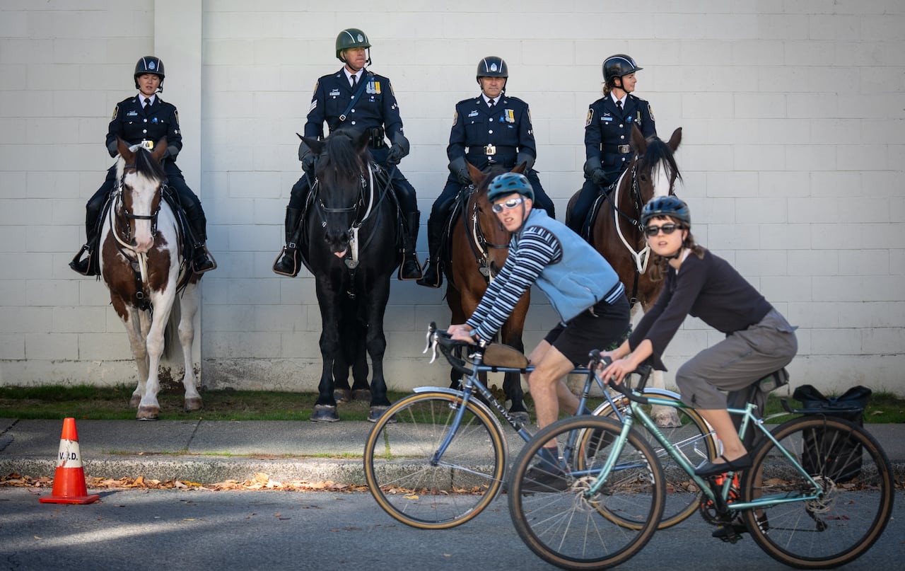Four police officers sit on horses against a wall as two cyclists bike by.