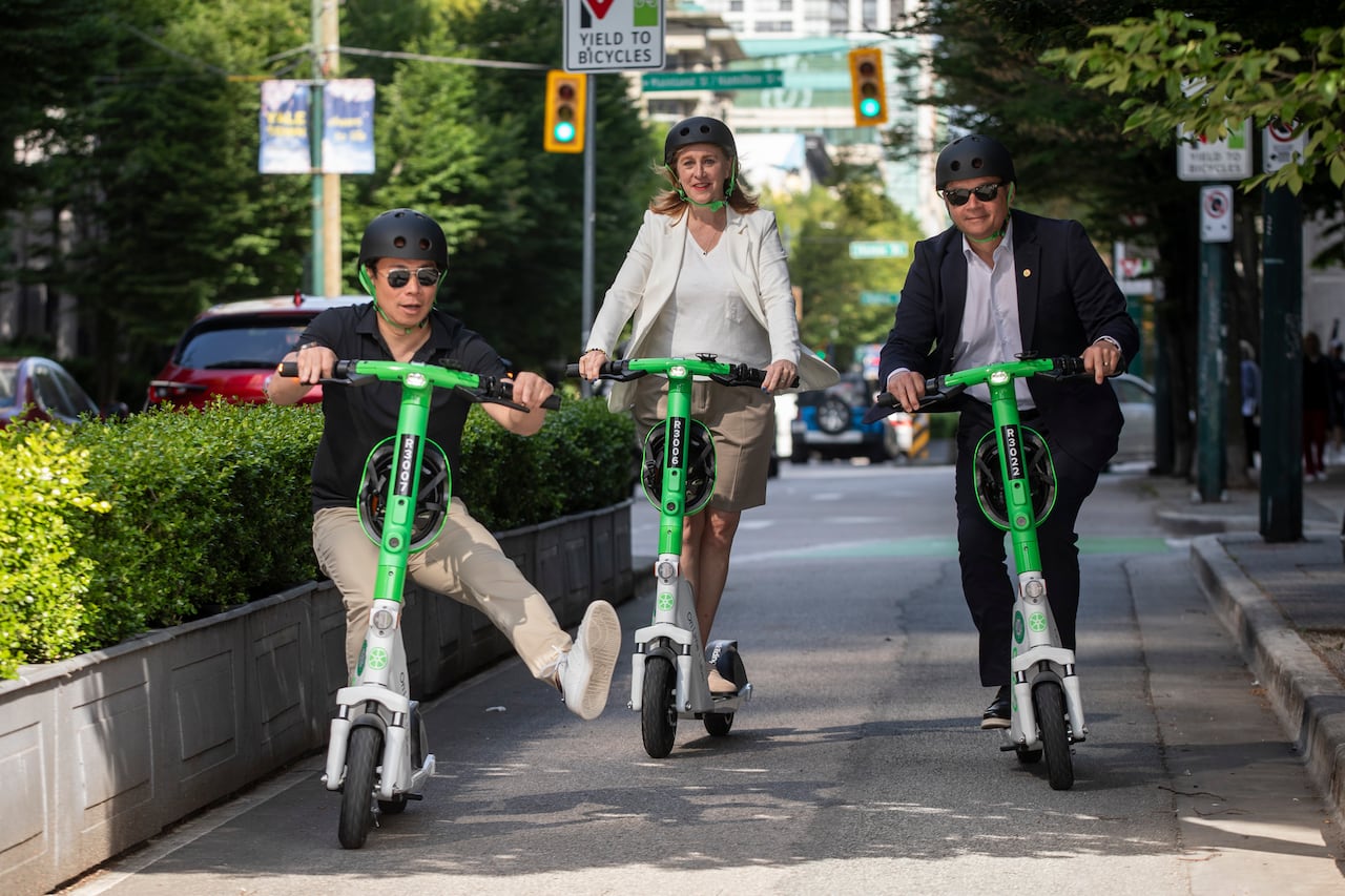 A man drives a scooter down a street while balancing on one leg with two others behind him.