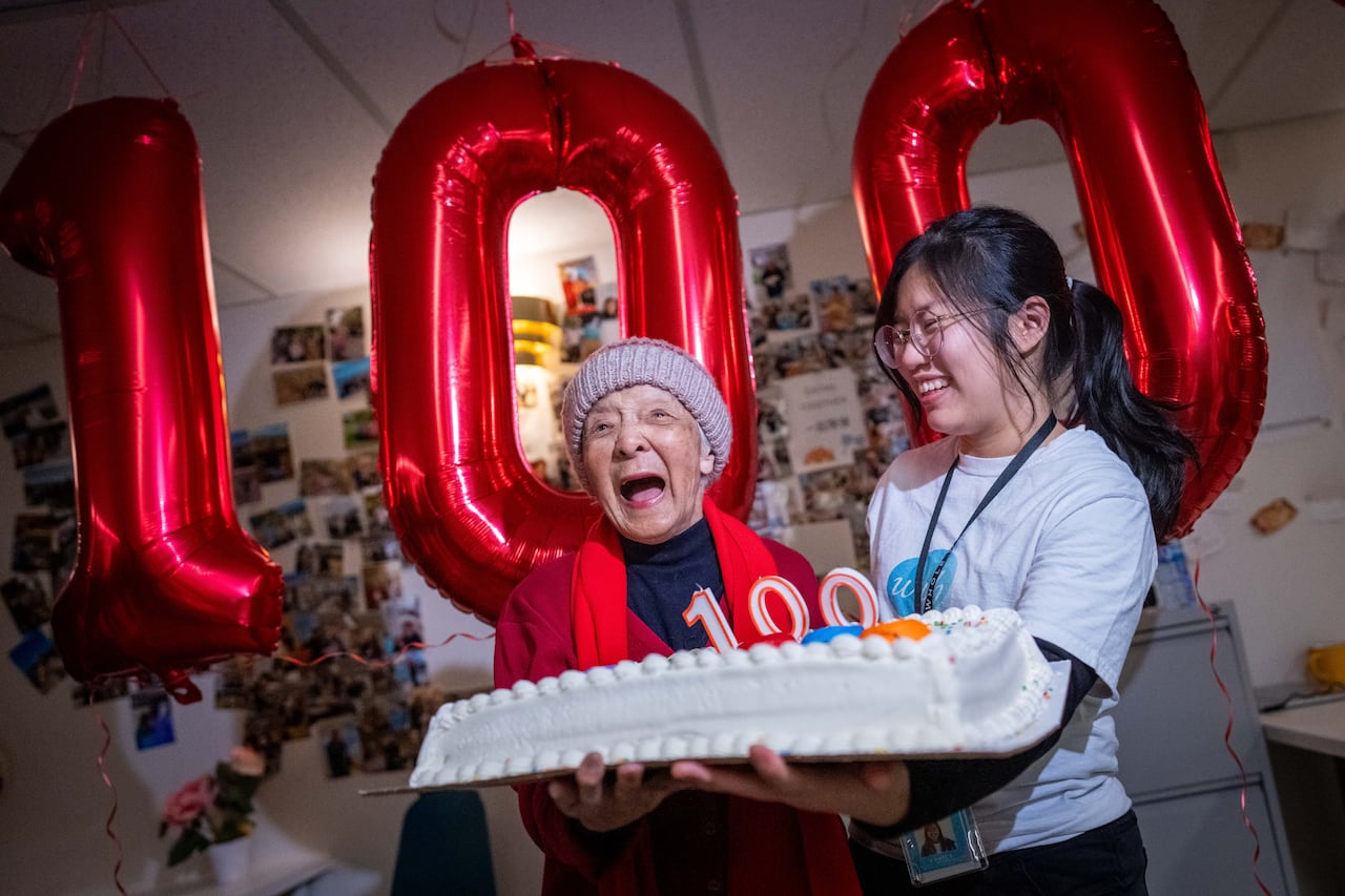 A woman holds a cake while laughing with a large balloon that says the number 100.