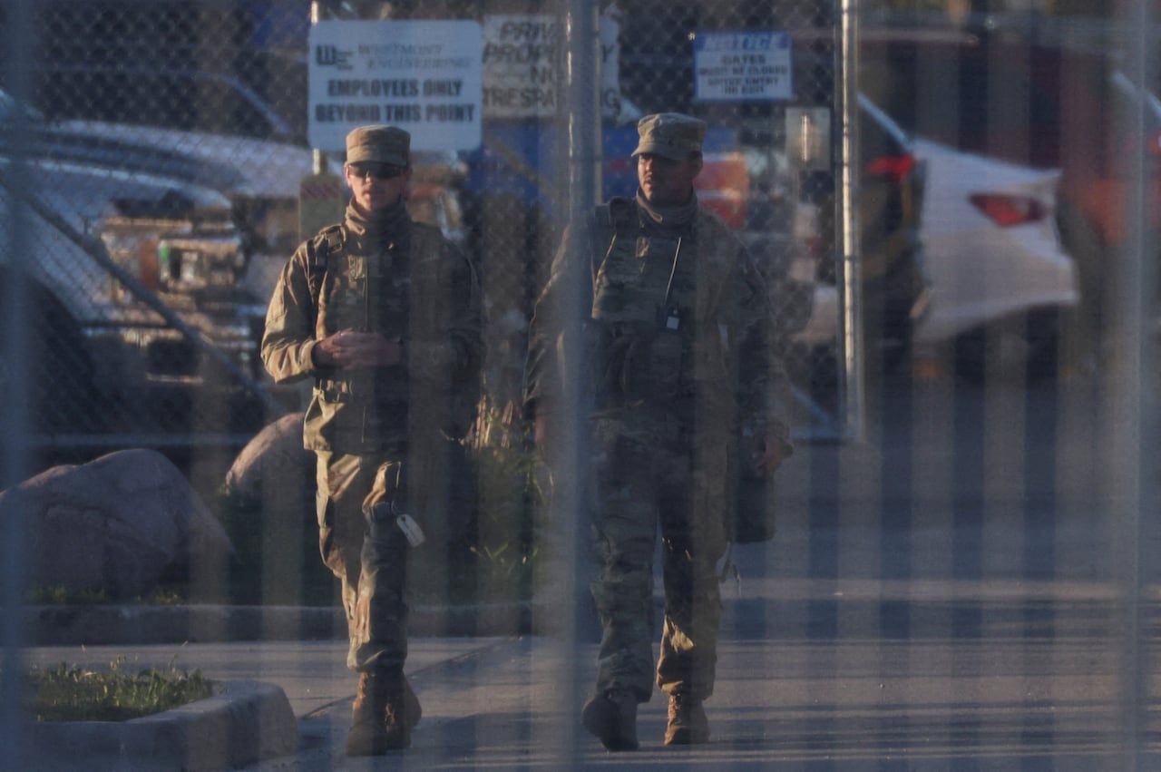 A view through a fence shows soldiers walking.