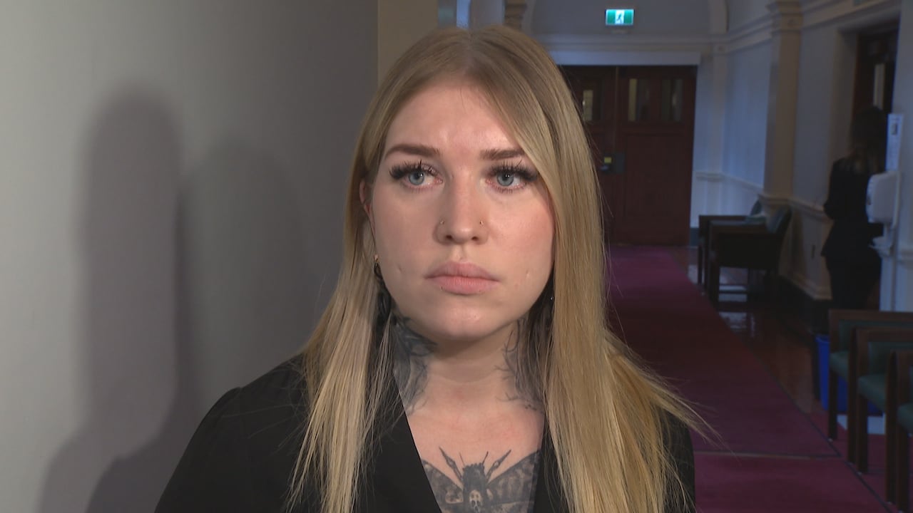 An woman with blond hair and tattoos on her neck is seen in a carpeted hallway.