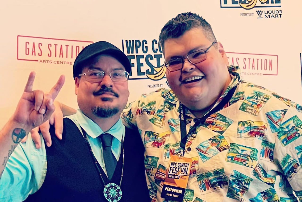 Two people posing for  photo. One is doing a devils horn sign, the other, who is wearing a Hawaiian shirt, is smiling.