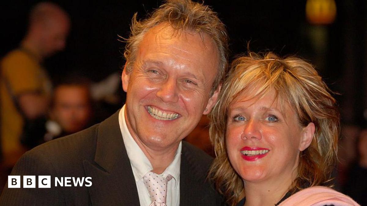 Anthony Head and Sarah Fisher attending Collars and Coats Gala Ball to mark the 150th Anniversary of Battersea Dogs and Cats home in 2010. Sarah is wearing a dark velvet style cape and a white blouse and Anthony is wearing a black suit, white shirt and tie. They have a white dog with brown and black markings, wearing a dog jacket between them. The dog is looking down to the floor and Sarah and Anthony are looking at the camera smiling.