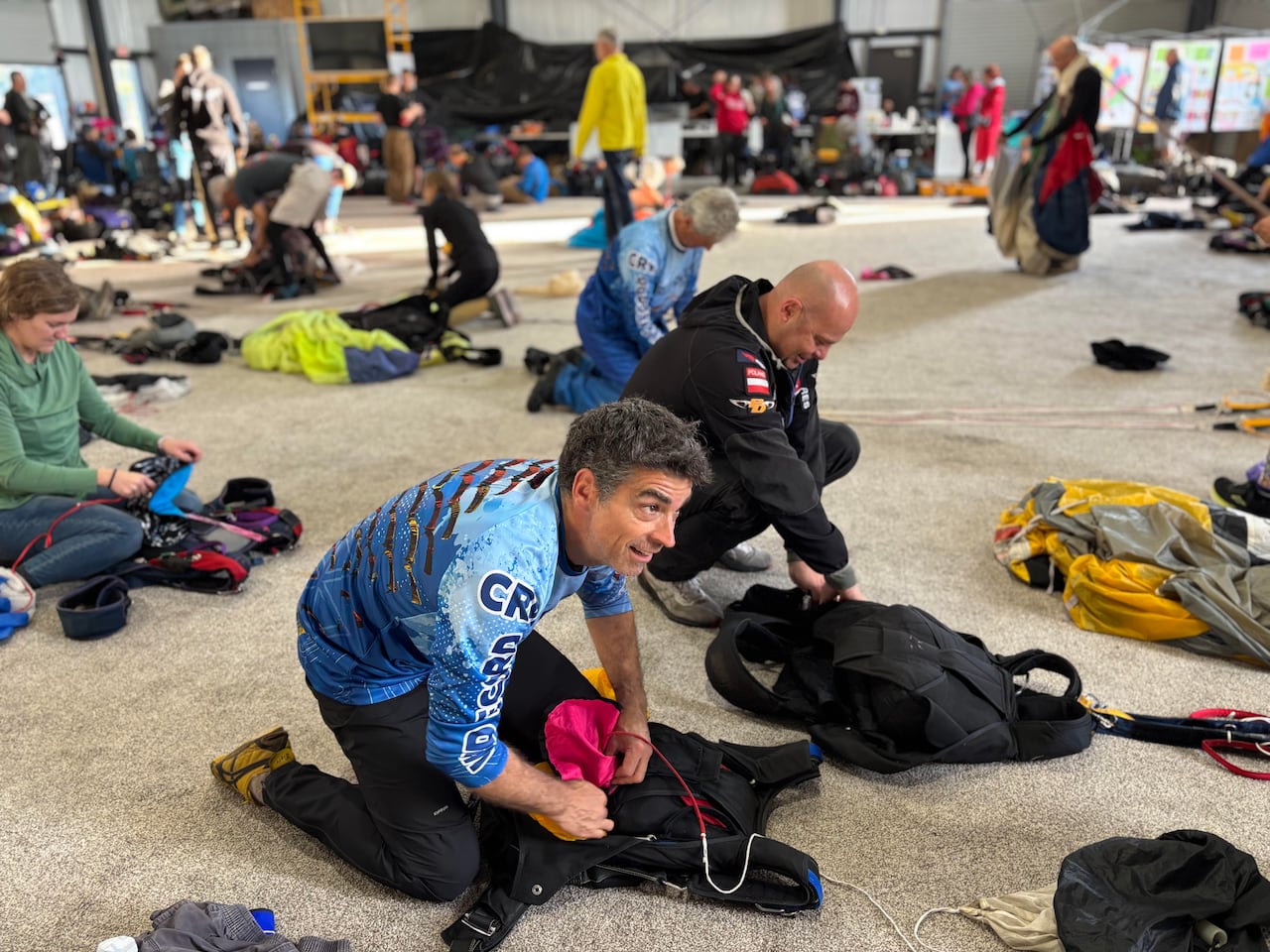 A group of people kneel on the ground and secure their parachutes into their packs.