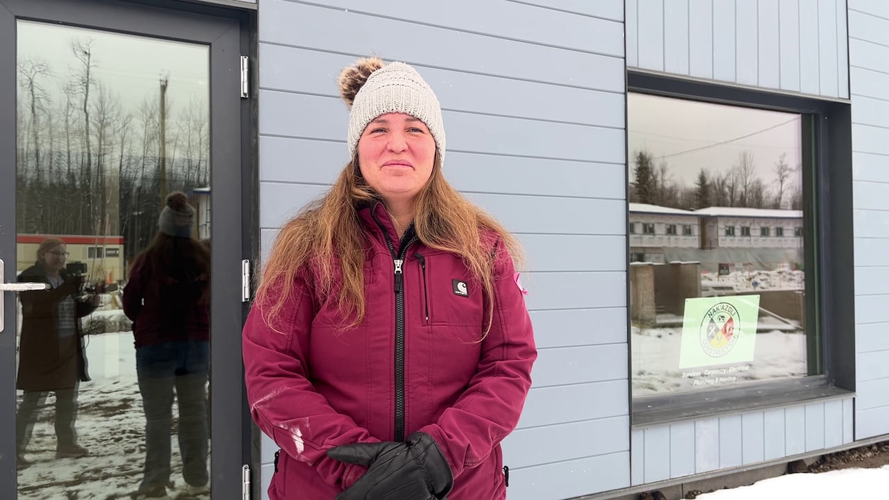 A woman in a pink jacket and white toque stands in front of a blue house. 