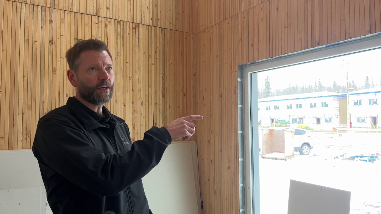 A man with a beard and short hair wearing a black jacket points to the wall inside a house with exposed wood on the walls. 