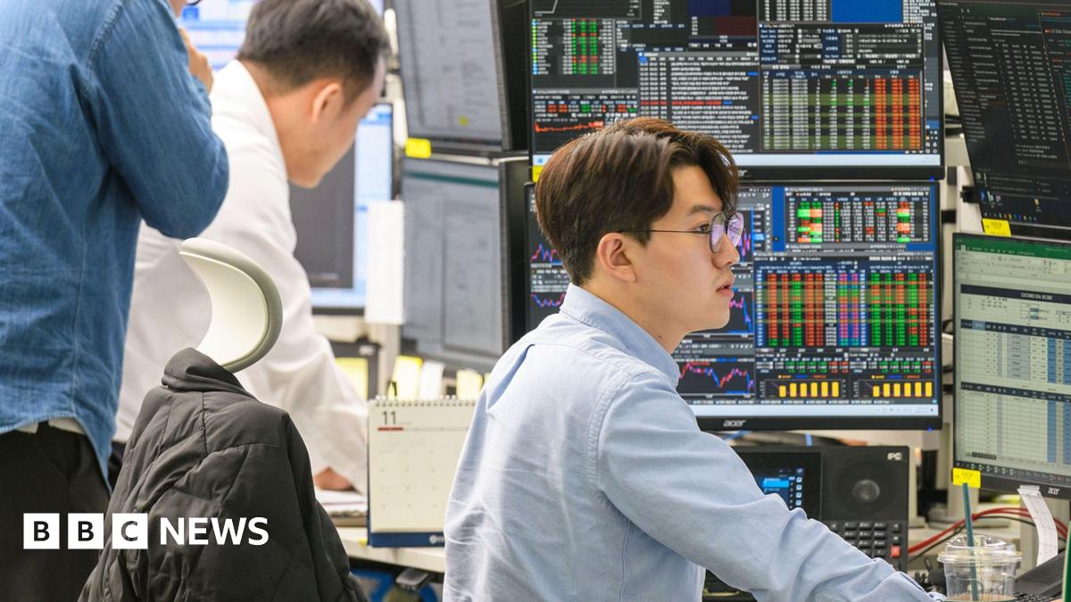 Three male city traders looking at multiple screens  with stocks and shares. Two large monitors show  financial graphs, charts, and data points in bright colors, including yellow, green, and red lines, indicating market trends. Image focused on the profile of one man