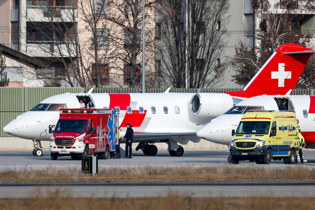 A white plane with the Swiss flag (a white cross on a red background) is parked on a tarmac with a red ambulance and a yellow emergency vehicle parked in front of it.