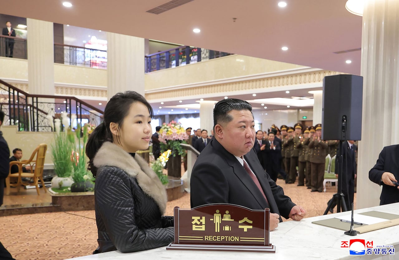 A girl in a black jacket with a fur collar stands next to an adult man in a black suit with a burgundy tie at a reception desk, a crowd of military officials behind clap behind them