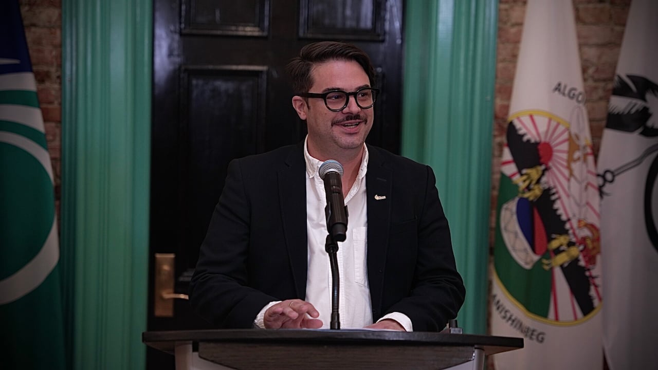 A man stands at a podium and makes a speech in a public building. A series of flags and a door are behind him.