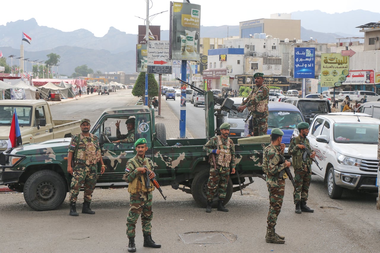 Men in camouflage fatigues and holding rifles stand in front a green military pick-up truck with another soldier standing on the back aiming a firearm that is mounted on the bed.