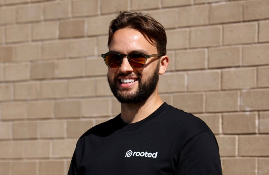 A man in sunglasses smiles in front of a brick wall.