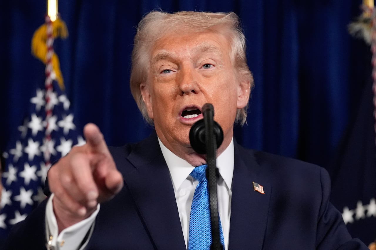 Man in navy suit and blue tie speaks into microphone with finger pointed