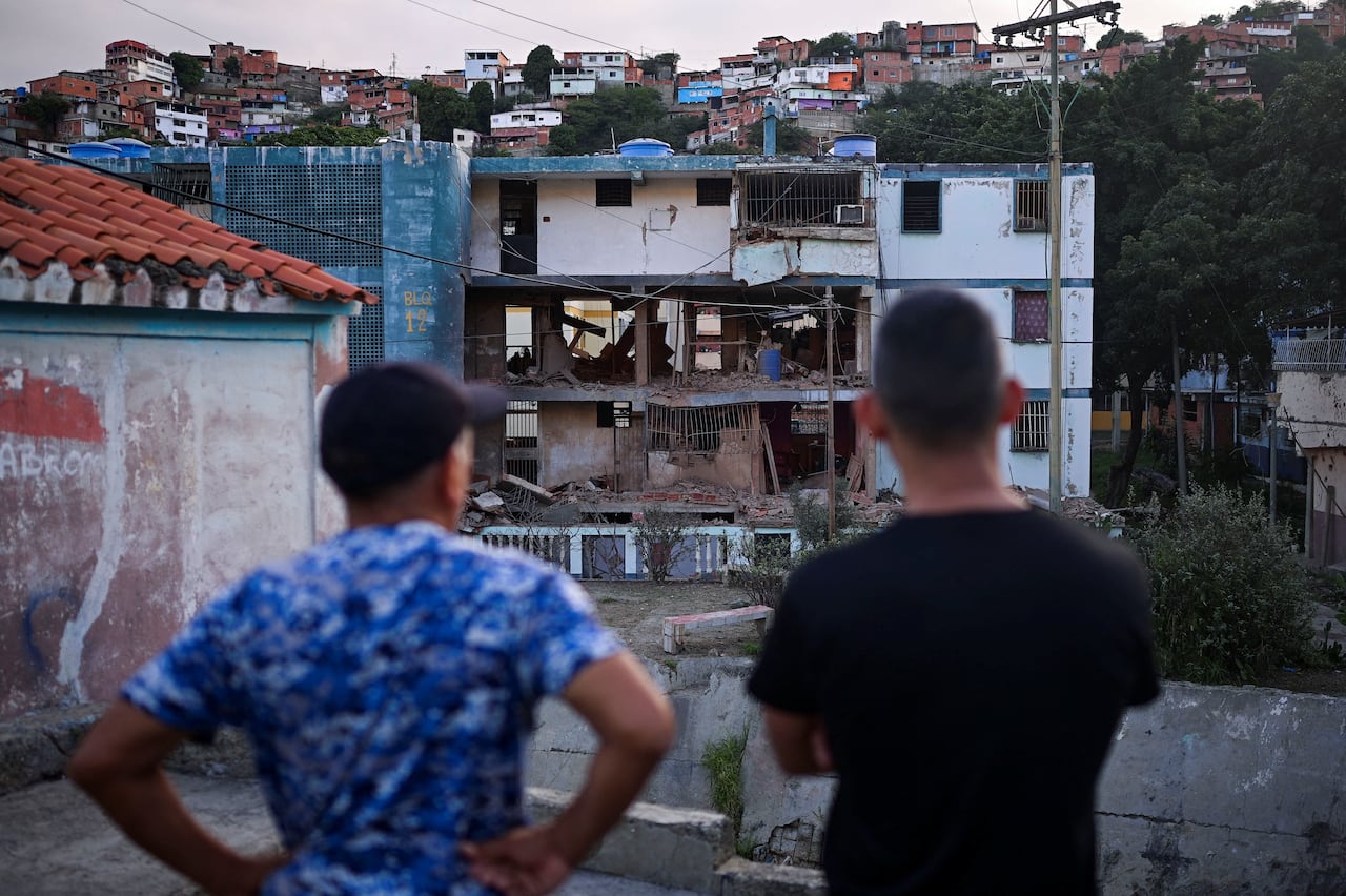 Two men observe a damaged residential building from afar.