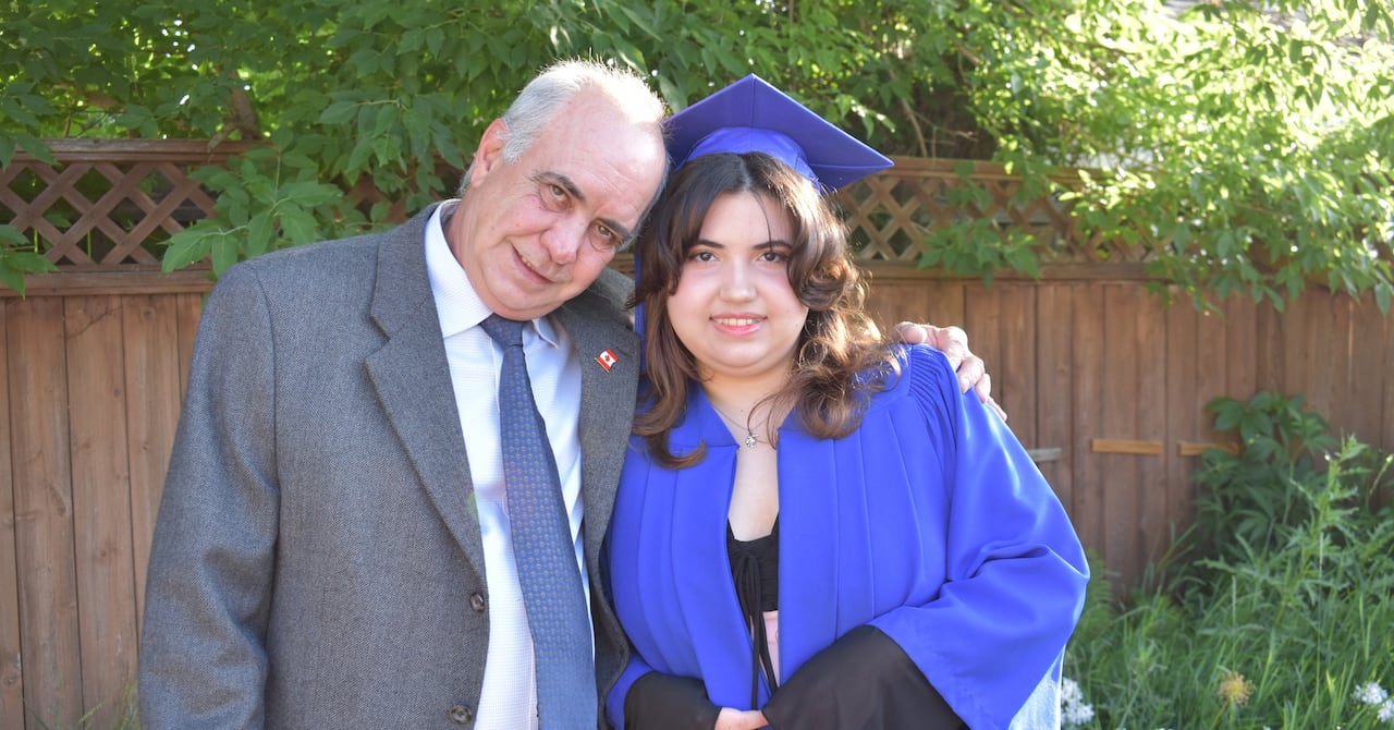 Man with white hair wears a grey suit and puts his arm around a young woman wearing a blue graduation gown and mortarboard cap.
