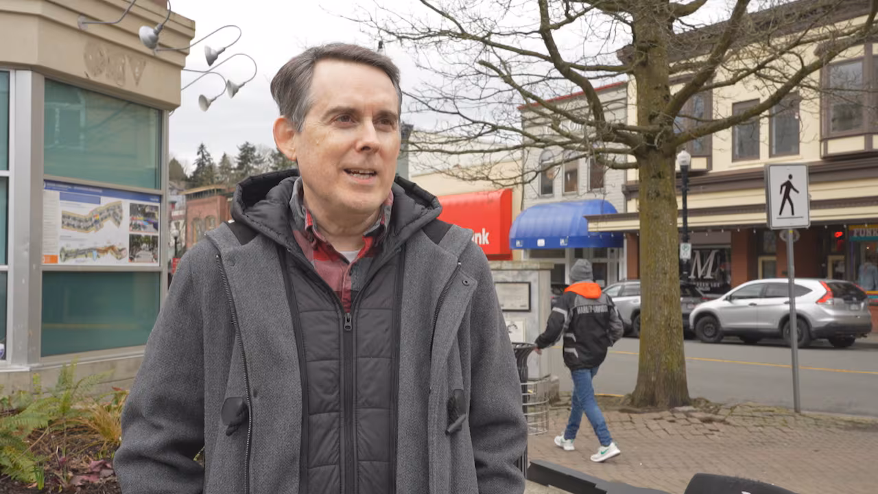 A man with short grey hair stands outdoors in a grey jacket. 