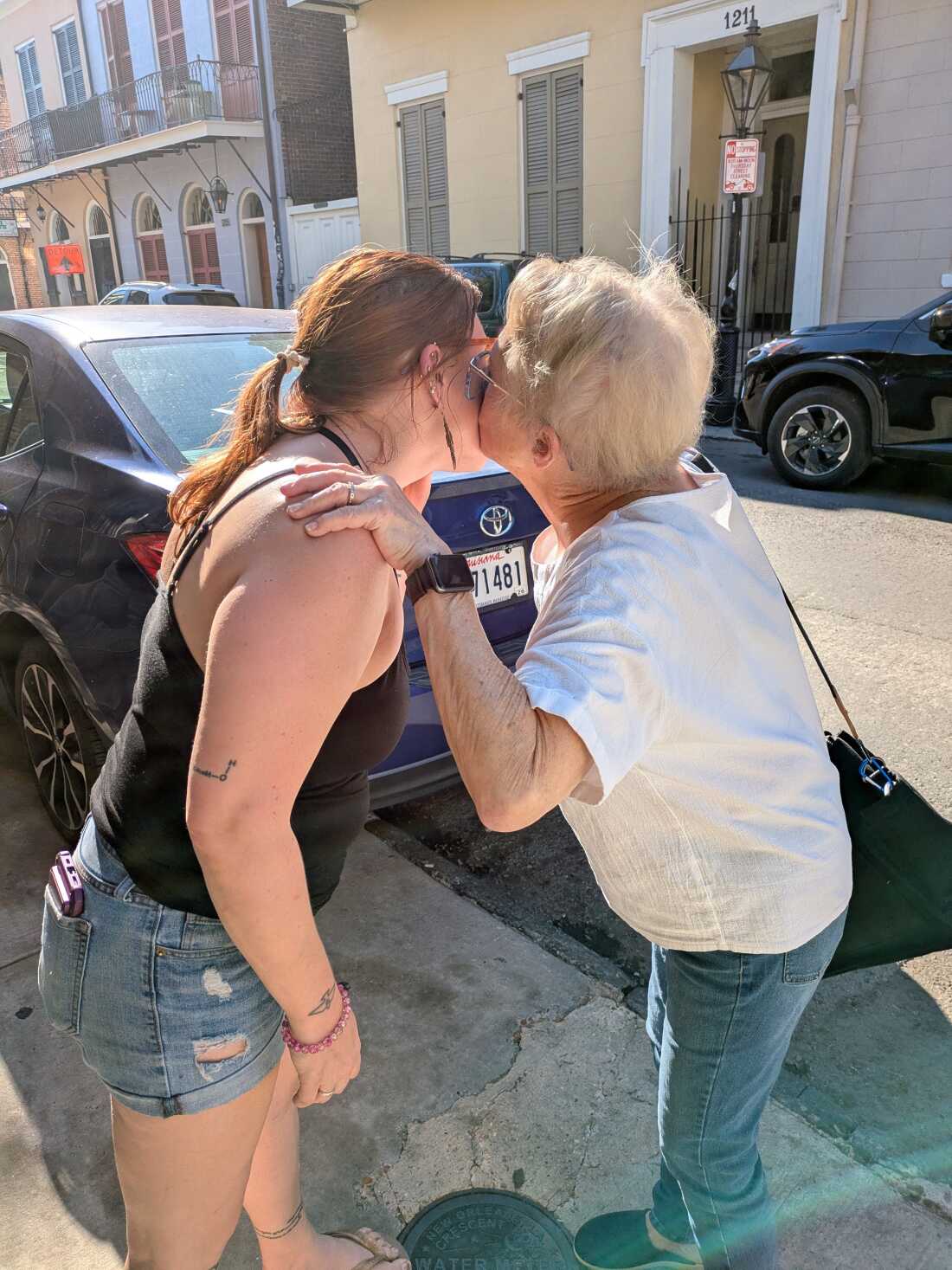 Dr. Elyse Stevens, who is wearing shorts and a tank top, gets a kiss from her mom, Mary Chaput. They are standing near a parked car on a street in New Orleans.