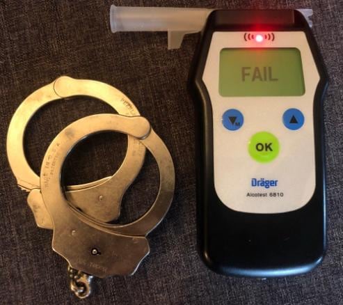 A pair of handcuffs lie on a table next to an alcohol breathalyzer that reads 'fail' on the screen.