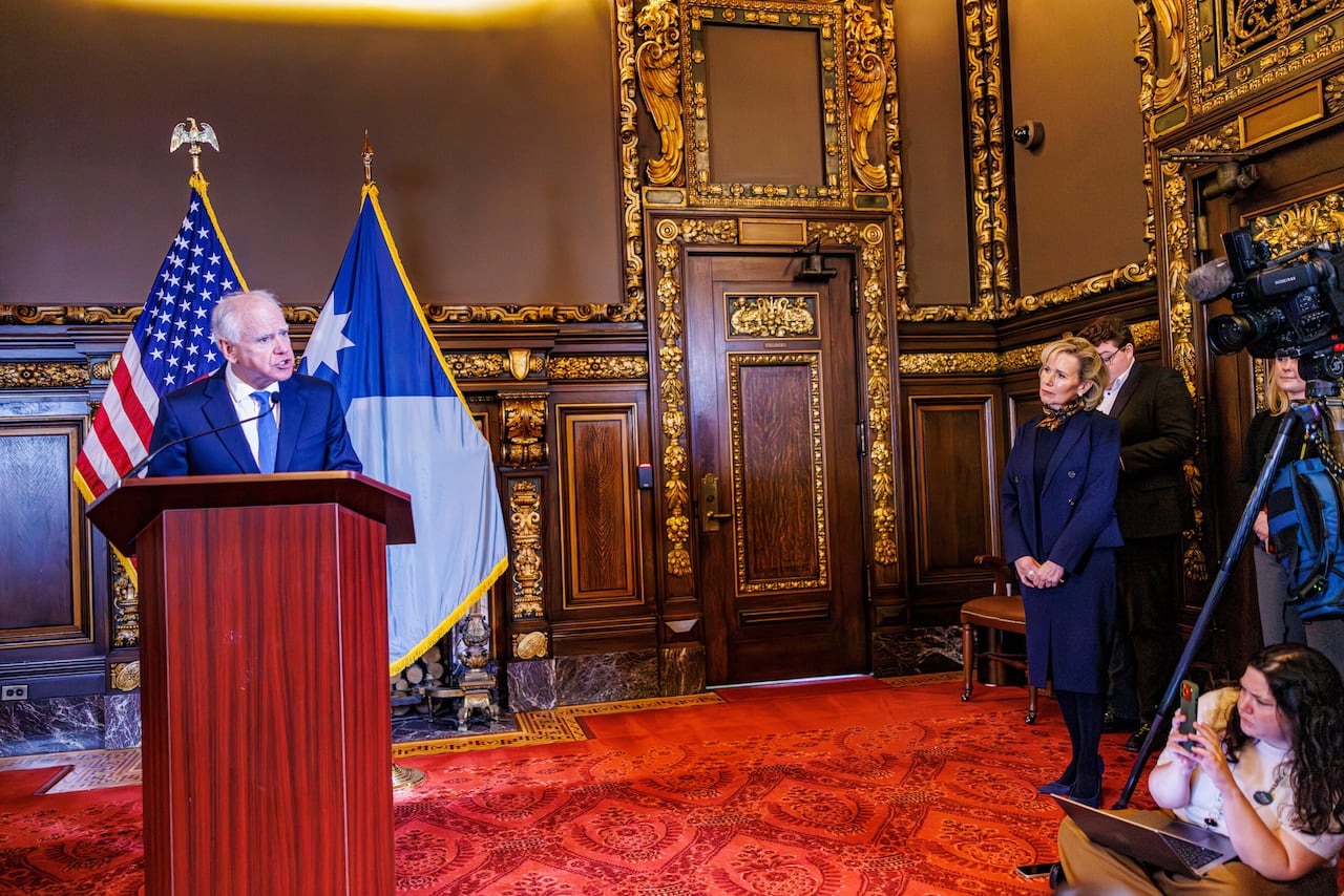 A person speaks while standing at a podium as other people and camera operators and sit stand nearby.