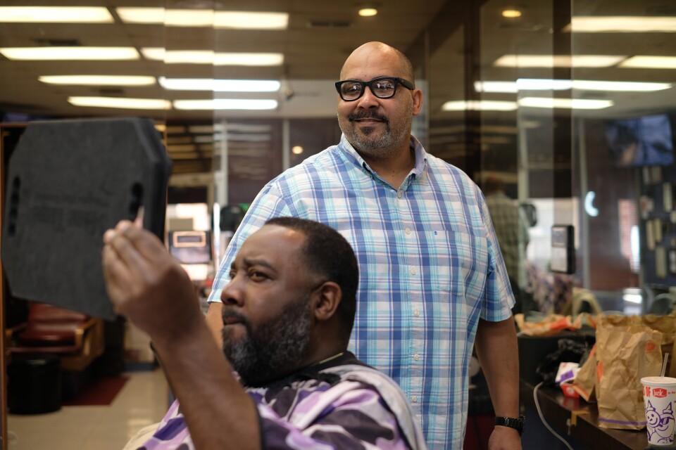 A man holds up a mirror to look at his haircut while barber Geoff Cathcart smiles behind him.
