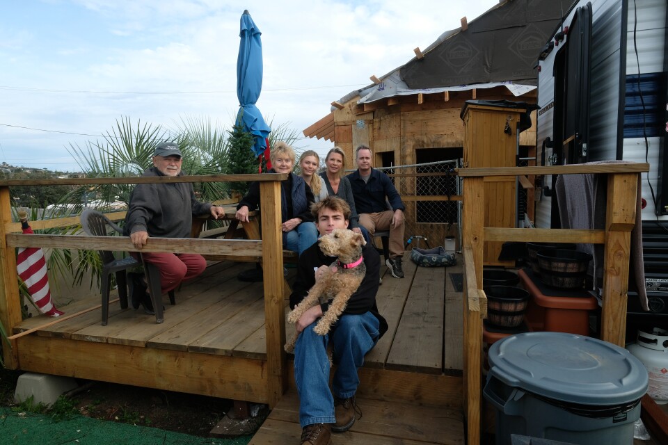 Three generations of a family sit on a deck of a home under construction. A young man sits on a step holding a brown dog.