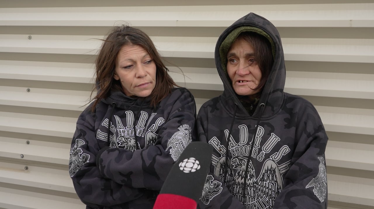 A woman with her hood up speaks to a mic, while another woman looks on.