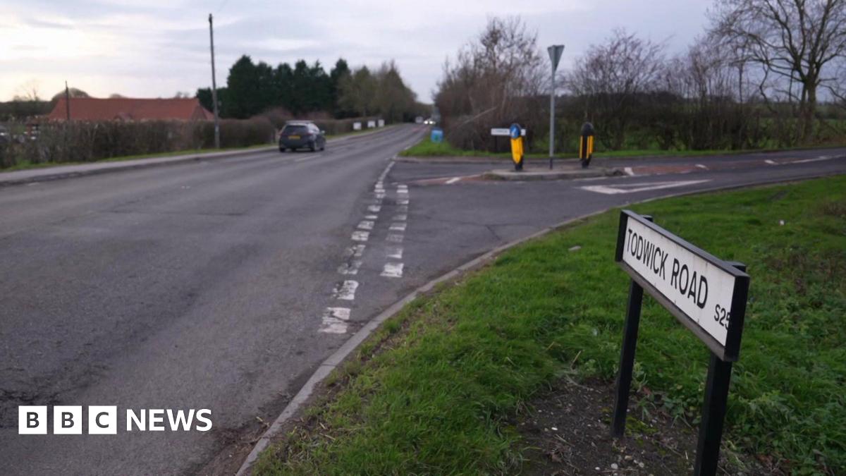 A rural road bordered by grassy verges and leafless trees. A black car is driving away in the distance. On the right side of the image, a white street sign reads “TODWICK ROAD S26” near a junction with traffic islands and bollards.