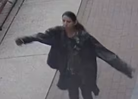 A grainy photograph of a woman wearing an over-sized black coat. She has dark black hair. Her arms are outstretched as she walks on a pathway.