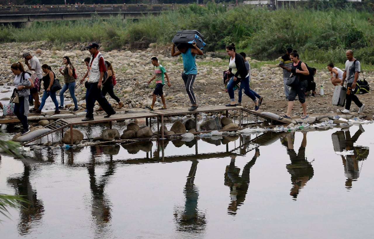 A line of people, some carrying bags in their hands and on their heads, walking over a small wooden bridge across a narrow river.