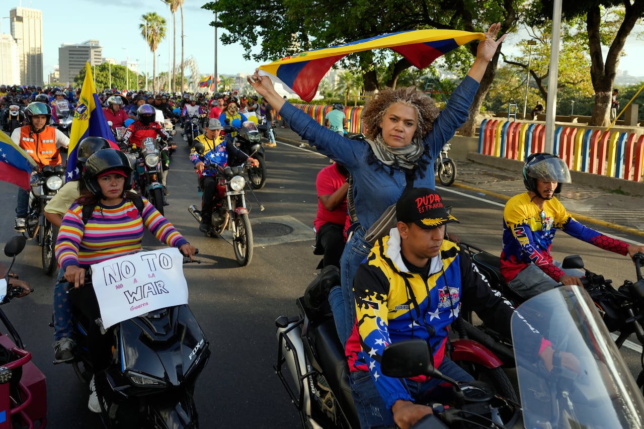 A woman sits behind a man on a motorbike holding a yellow, blue and red Venezuelan flag behind her head as other people on motorbikes follow along a road.