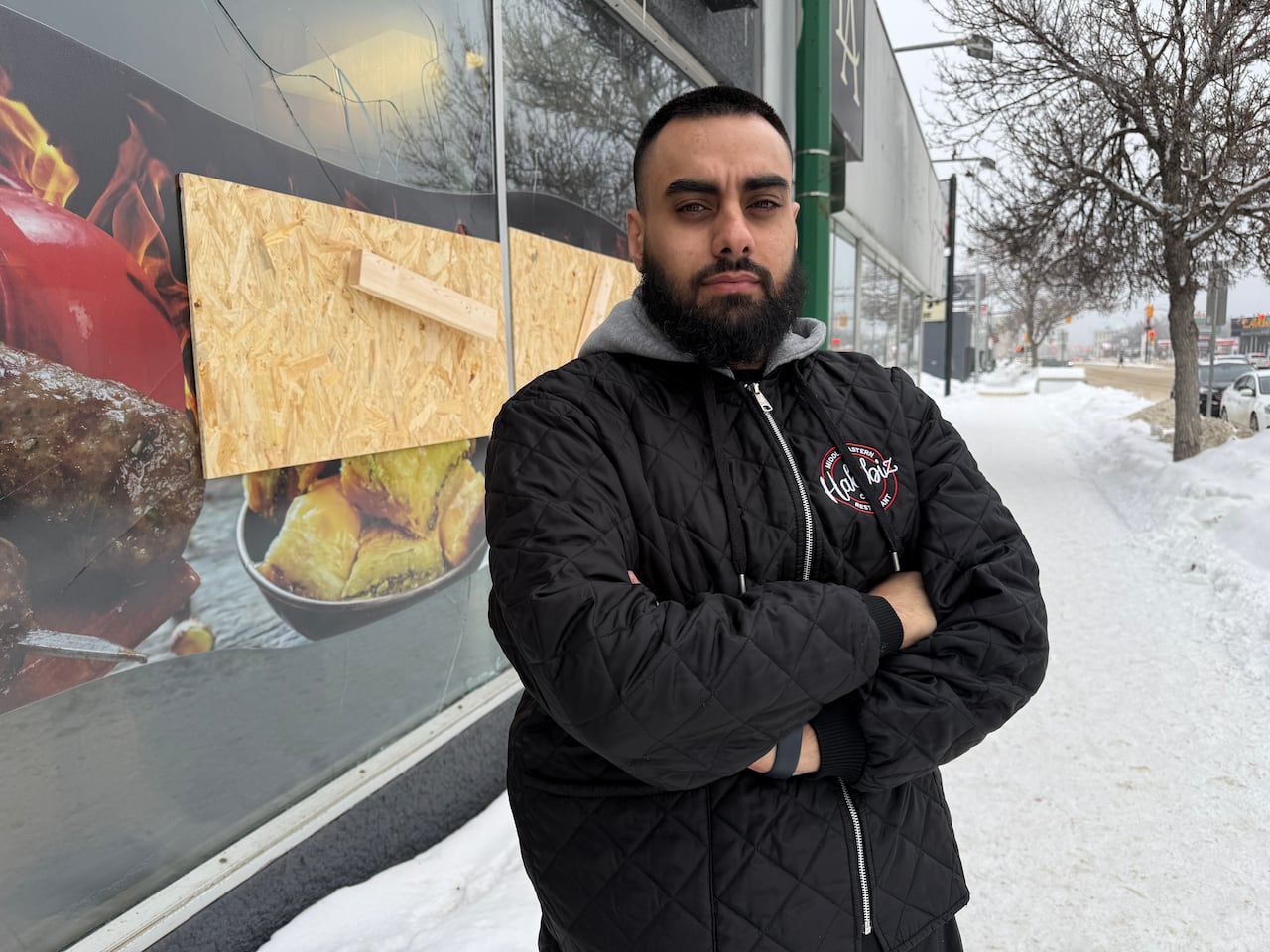 Am man in a black jacket with his arms crossed stands outdoors in front of a broken window boarded up.