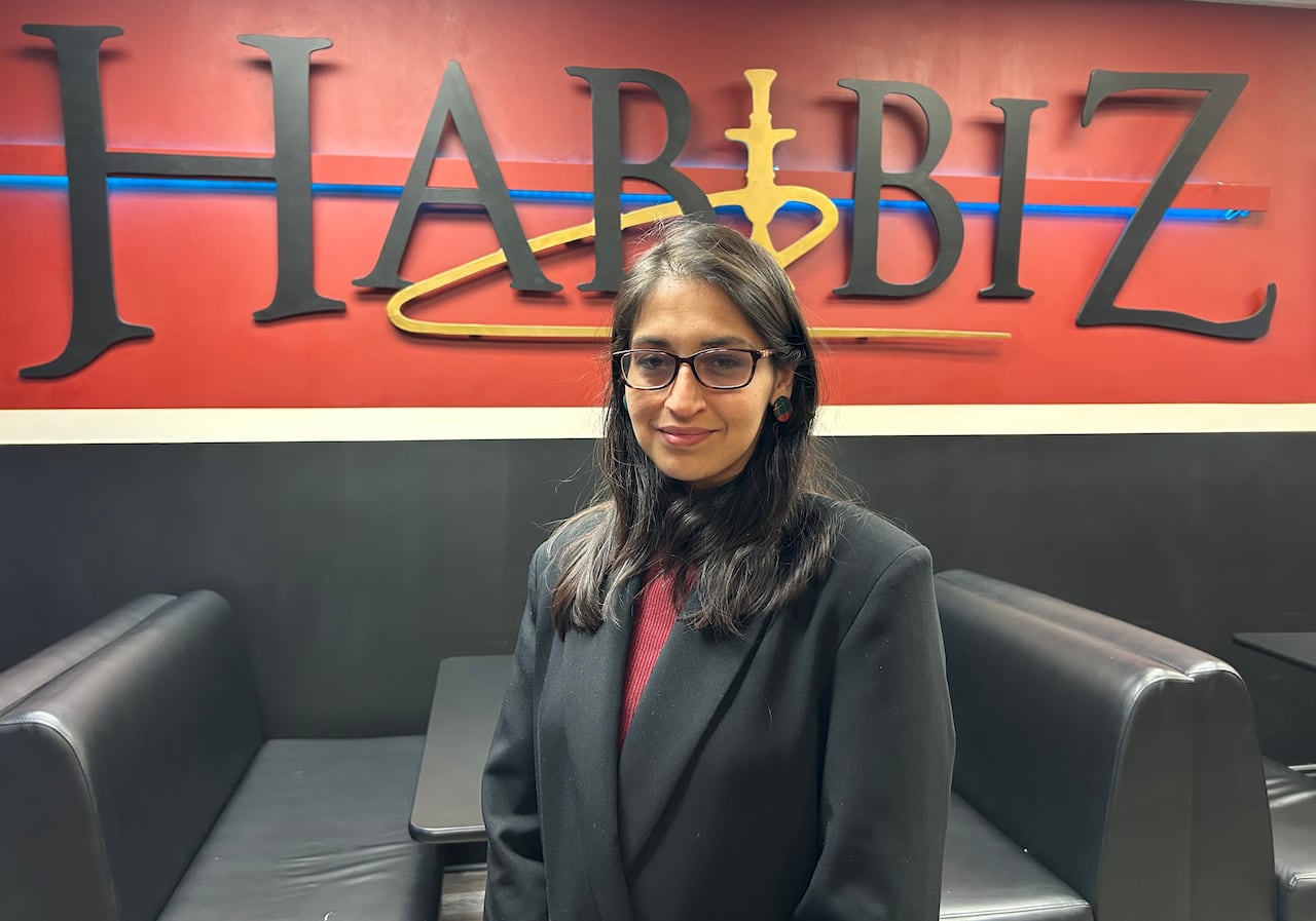 A woman with long dark hair, wearing a dark blazer and red shirt, stands in front of a sign that reads 'Habibiz.'