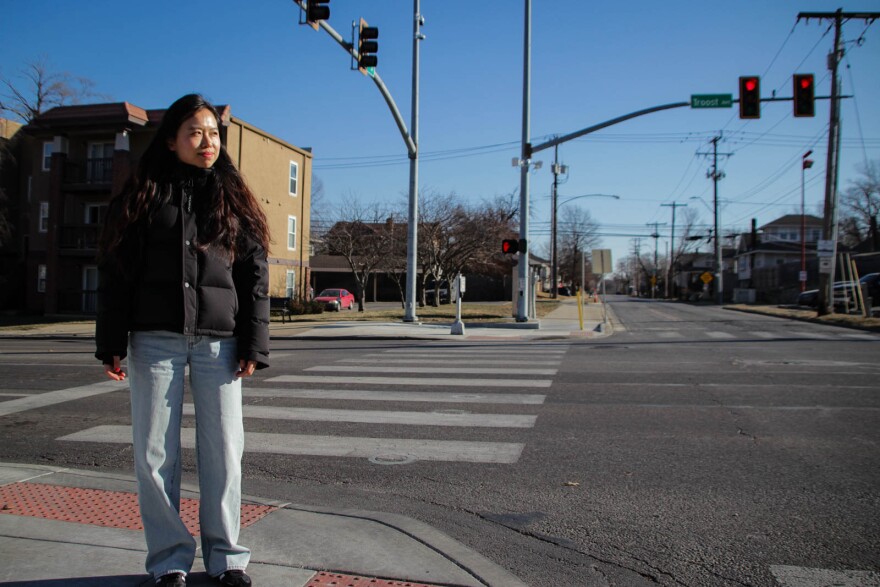 A woman in jeans and a black puffer jacket stands on a sidewalk looking towards her left.