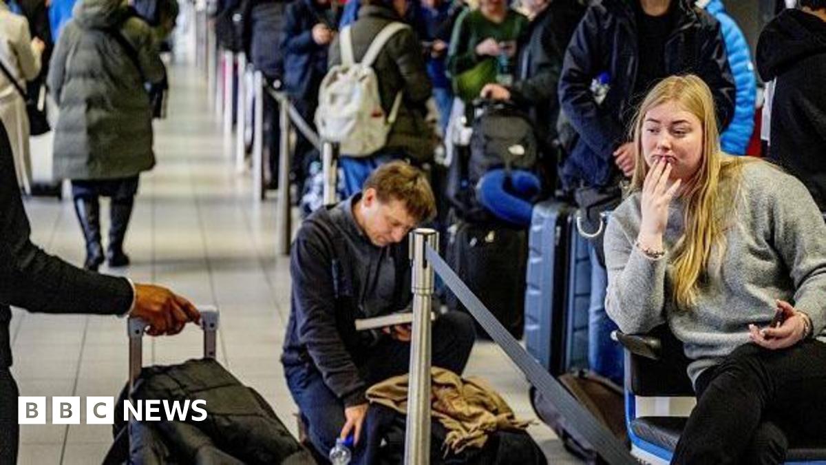 Travelers queue with their luggage at Schiphol Airport. One woman is sitting with her phone in her hand, while a man behind her is kneeling next to his bag while reading a book.