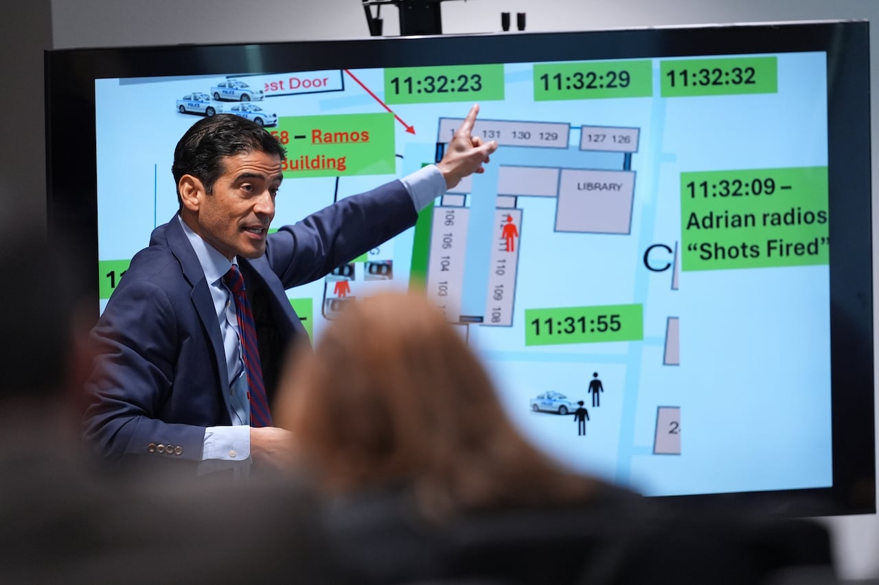 Dark haired man in a blue suit gestures to a poster board as he speaks