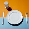 This image shows a high-angle view of a white plate, a stemmed drinking glass, a fork to the plate's right and a knife to the plate's left. The items are all set on a tabletop that's orange across the top of the frame and light blue across the bottom.