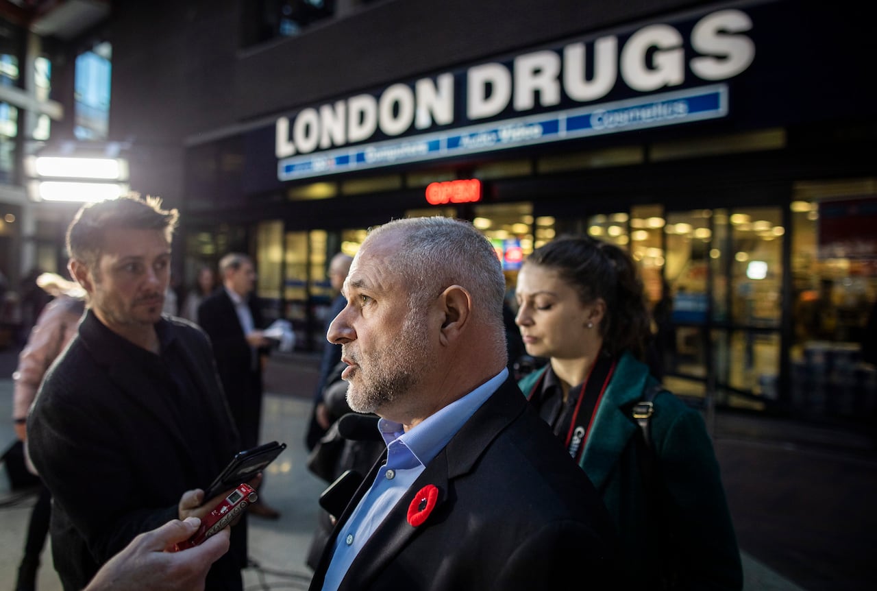 A balding white man is seen speaking to reporters outside a retail outlet that says 'London Drugs'.