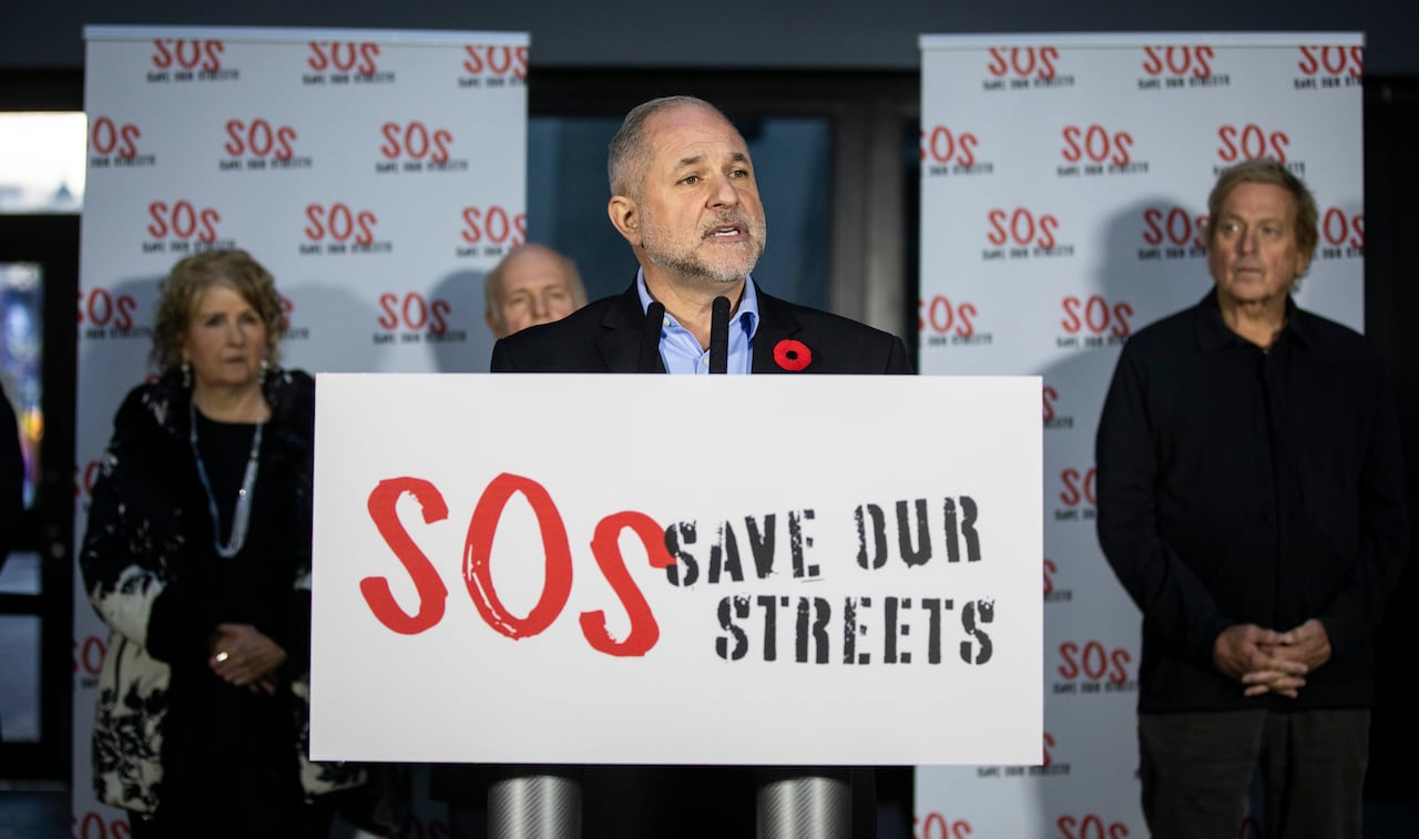 A balding white man, flanked by a few others, speaks at a podium marked 'Save our streets'.