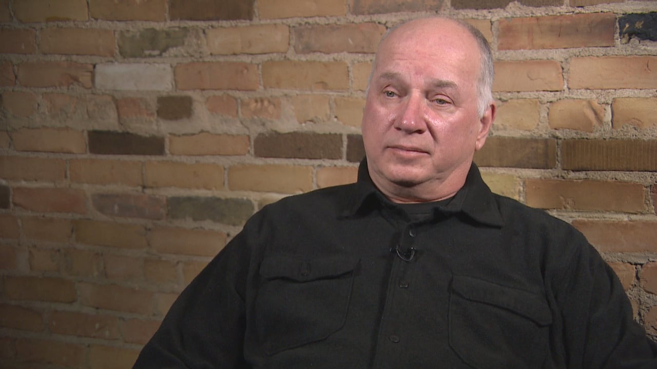 A man in a black shirt sits in a front of a brick wall.