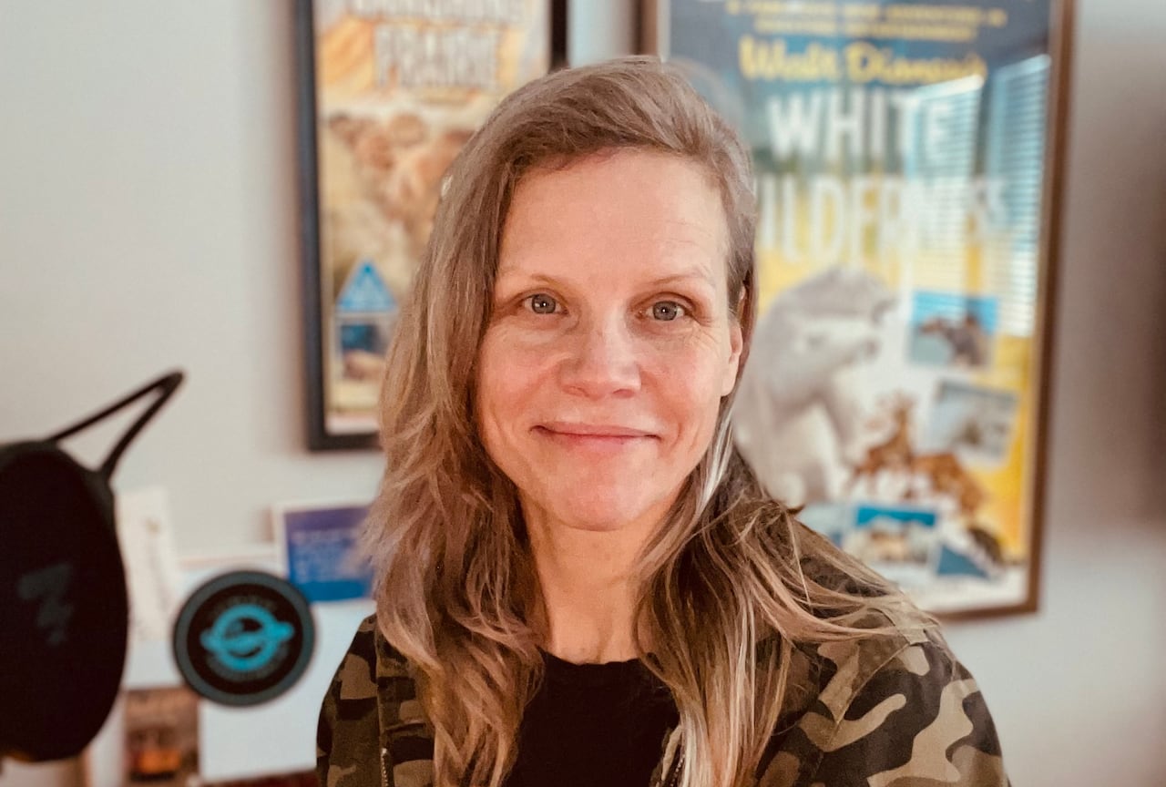A woman with blonde hair wearing a camo jacket and a Led Zeppelin shirt sits in a sunlit room with movie posters in the background.
