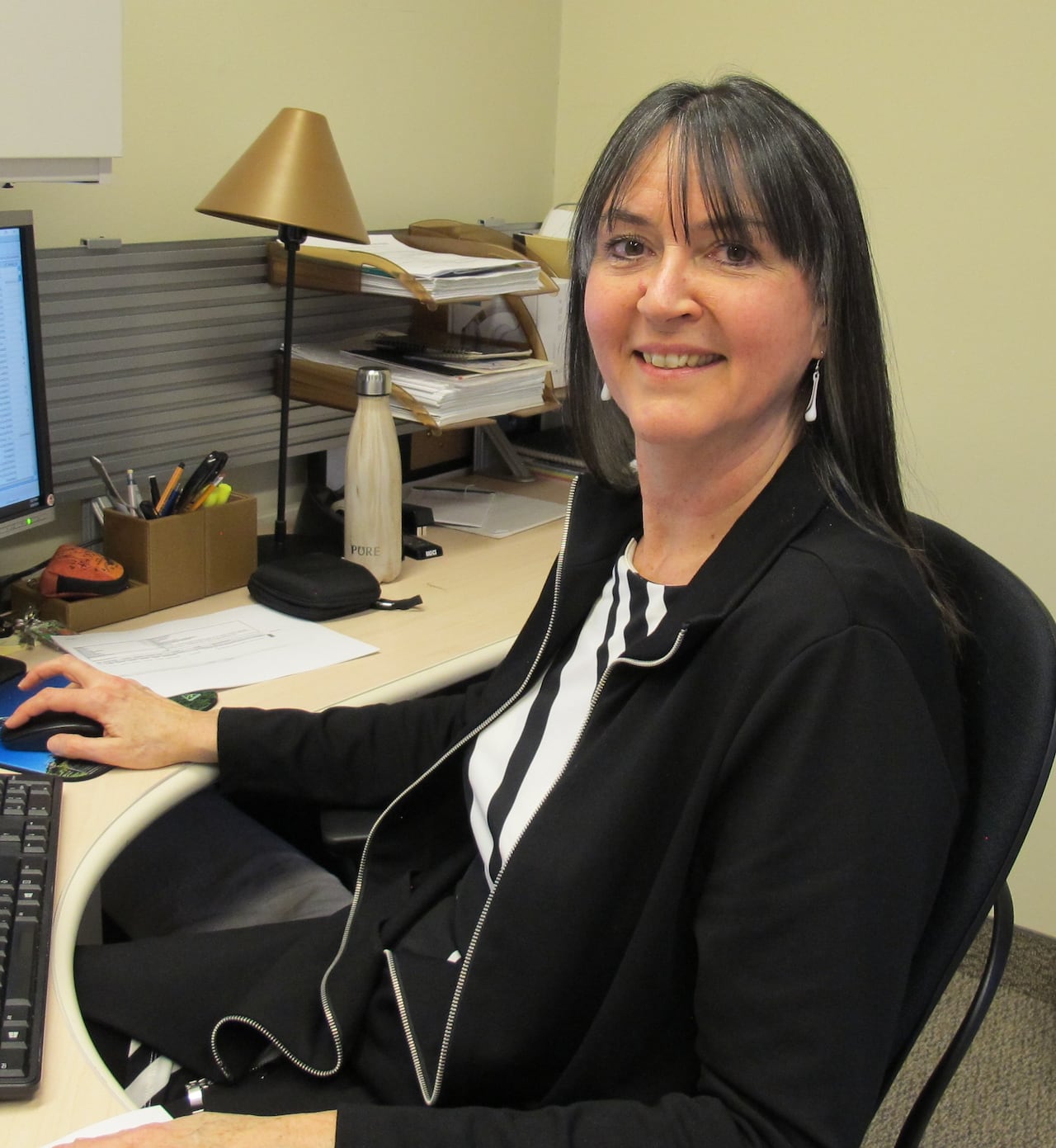 A woman sits at a desk in front of a computer. She is shown from the side and smiles with her head turned toward the camera. She has dark hair with some grey and is wearing black and white.