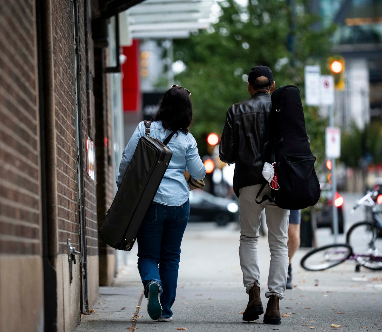 Two musicians carrying instruments in carrying cases are seen on a city street from behind.