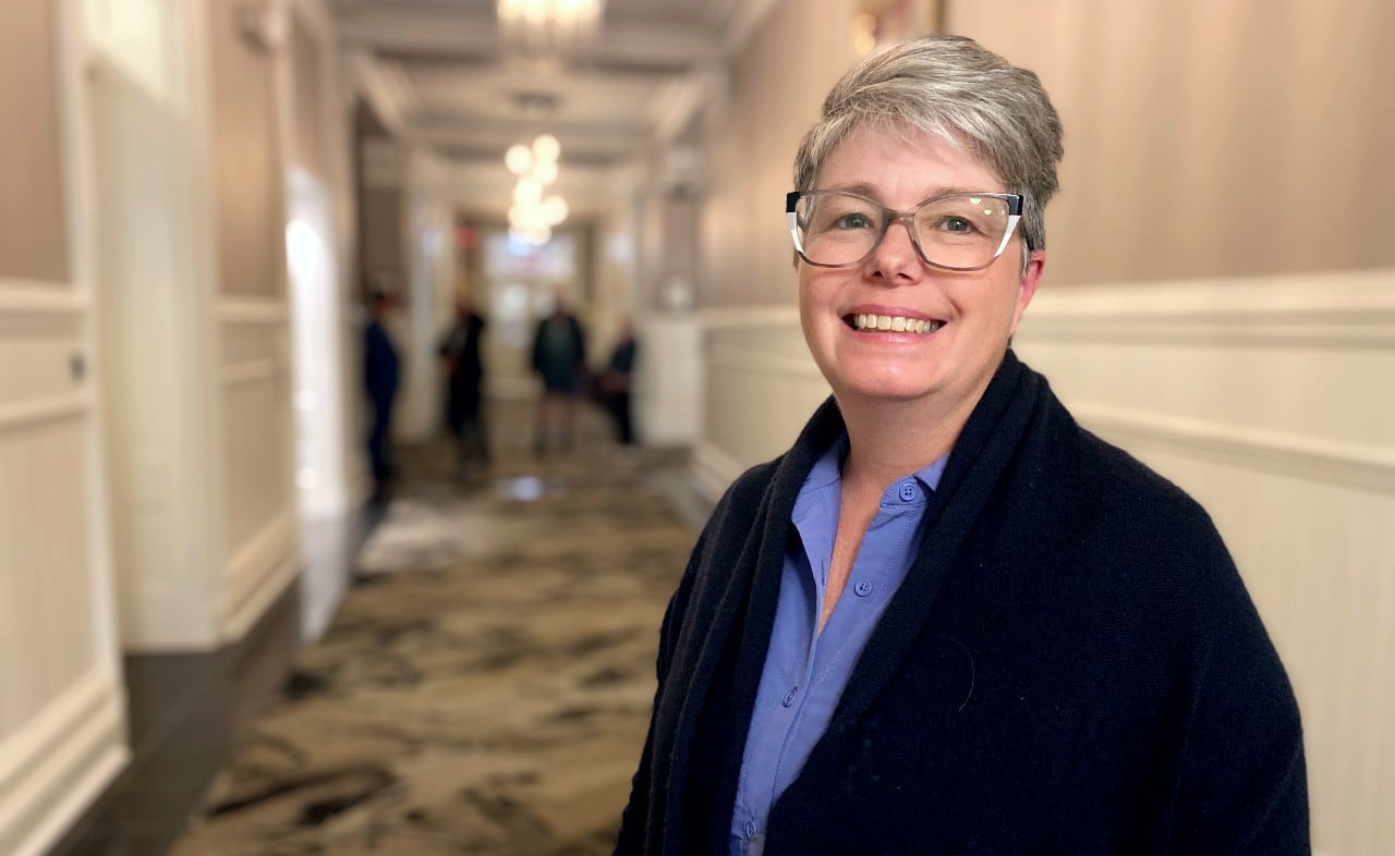 A white woman with short grey hair and glasses wears a blue shirt under a black cardigan. She stands in a beige carpeted hallway