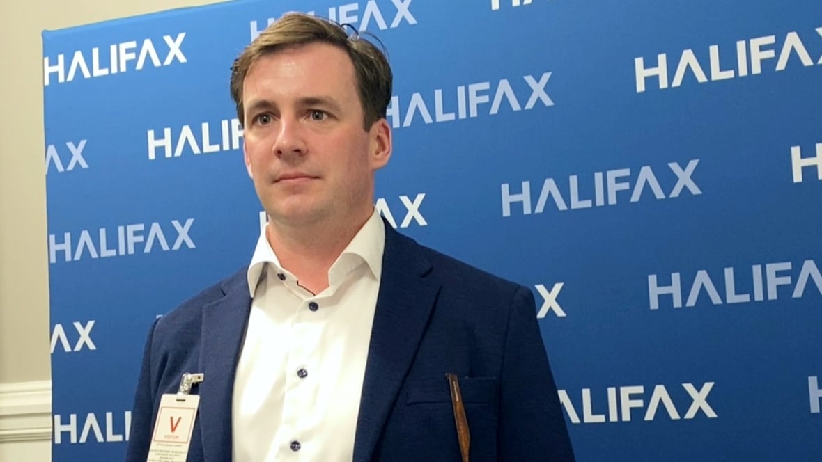A white man in a blue suit and white shirt stands in front of a blue backdrop with the Halifax city logo on it