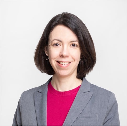 A woman with chin-length brown hair, and wearing a grey blazer and bright pink shirt, is smiling at the camera.