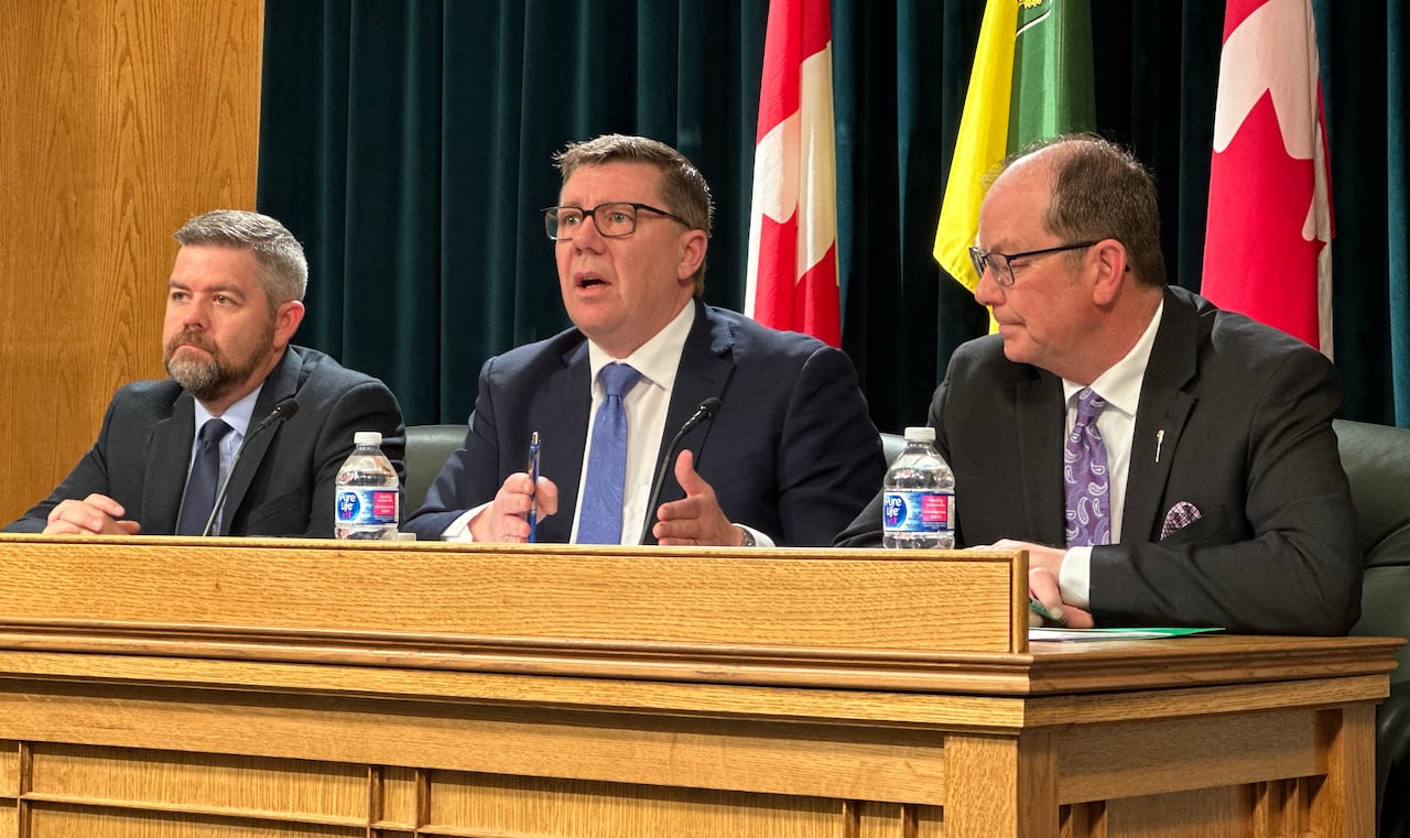 Three men in suits sitting at a wooden desk with flags in the background.